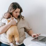 Young working mother cuddling baby and using laptop at home